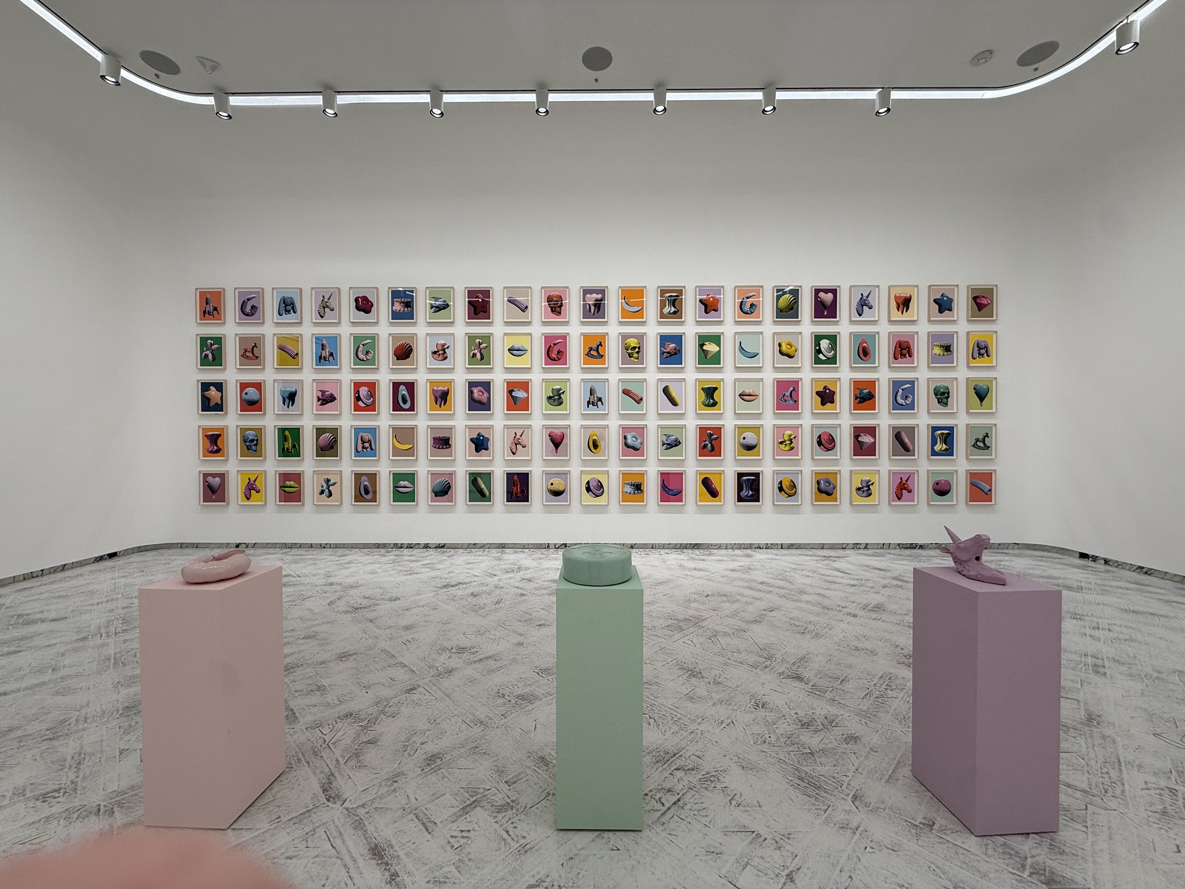 A wall of framed hyperrealistic bead paintings, sculptures on pedestals below